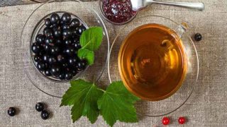 Black currant tea in a cup, next to blackcurrant fruits in a cup, and blackcurrant leaves on a table