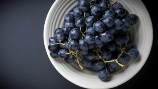 A white bowl of purple grapes on a dark counter