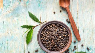 A bowl of black pepper corns with a wooden spoon and fresh leaves on a table