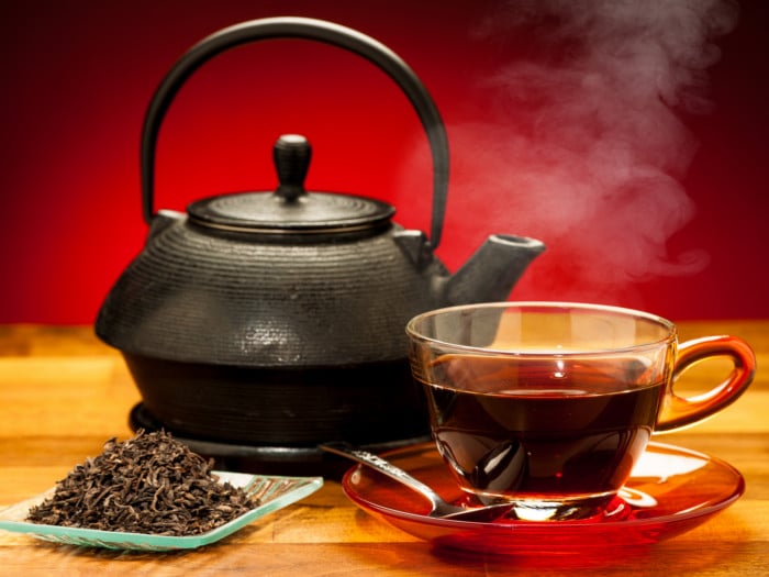 A cup of black tea, a black kettle and a plate of black tea leaves on a wooden table