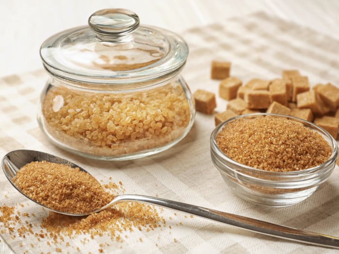A jar of granulated brown sugar, a spoon and bowl of brown sugar powder, and brown sugar cubes on a cloth