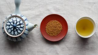 A flat lay picture of a kettle, buckwheat seeds, and buckwheat tea kept in line next to each other