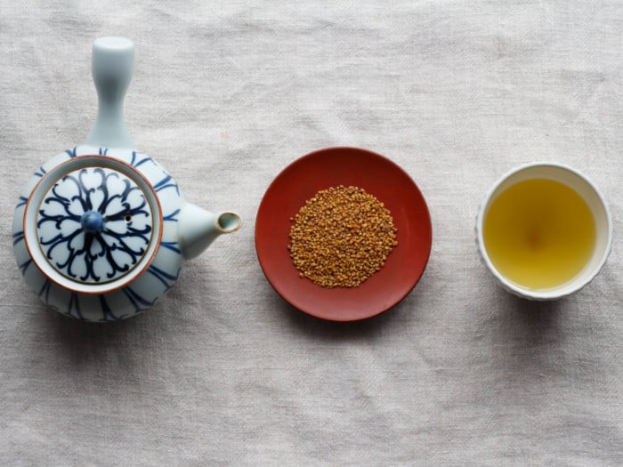 A flat lay picture of a kettle, buckwheat seeds, and buckwheat tea kept in line next to each other