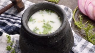 Black earthenware pot, wood churner, sliced onion, and coriander sprigs, placed on a white and blue check cloth.
