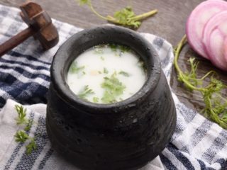 Black earthenware pot, wood churner, sliced onion, and coriander sprigs, placed on a white and blue check cloth. 