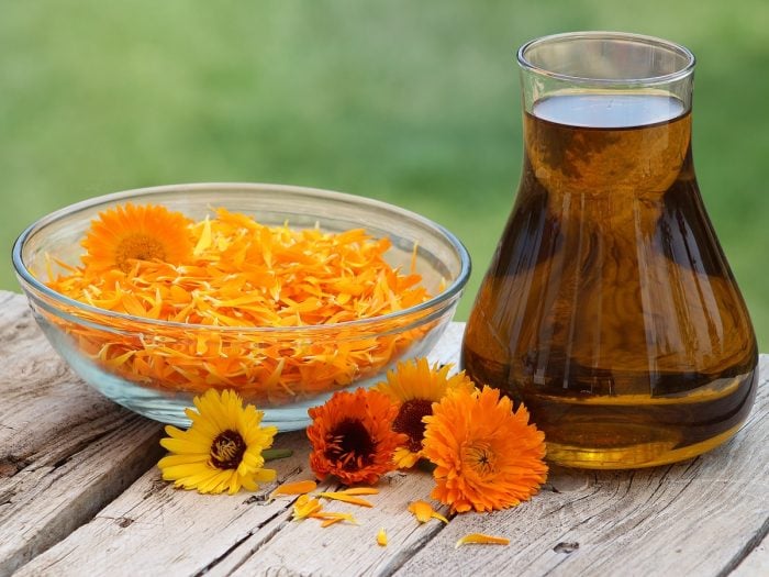 Calendula oil in a jar placed next to calendula flowers on a wooden platform against the background of green grass