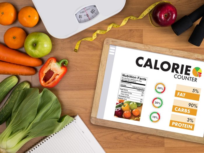 A weighing machine, measuring tape, and a slate with a calories counter on it surrounded by vegetables atop a table