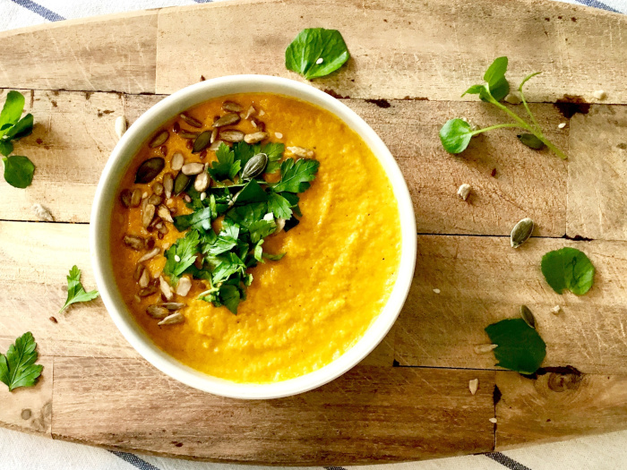 Carrot soup with coriander, parsley, and seeds in a white bowl on a wooden board.