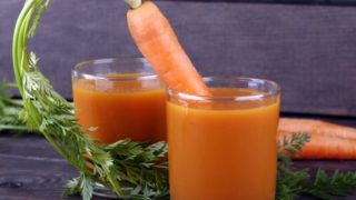 A carrot sticking out of one of the two glasses of carrot juice placed atop a black table