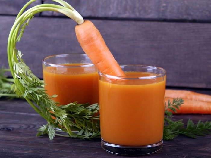 A carrot sticking out of one of the two glasses of carrot juice placed atop a black table