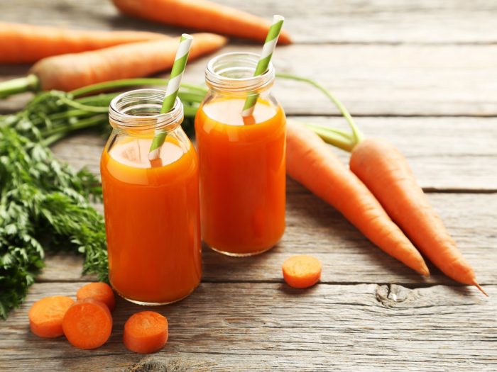 Two bottles of fresh carrot juice with straws inside, whole and halved carrots and cilantro leaves on a wooden table