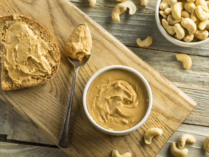 Cashew butter in a white bowl and on a bread slice with cashews in the background