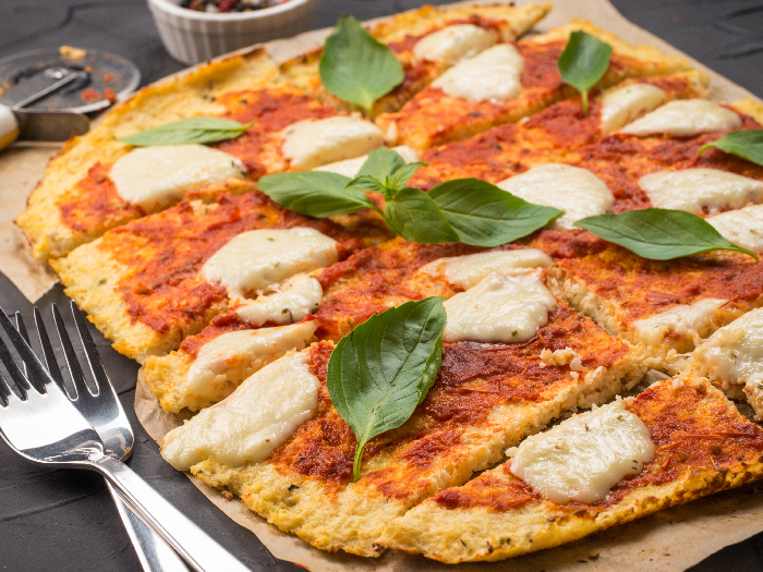 sliced homemade cauliflower pizza crust with mozarella and fresh basil in dripping pan on black concrete background