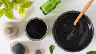 Flatline view of a bowl of black viscous liquid with a wooden spoon, smaller bowl of black powder, different-shaped stones, and leaves on a white background.