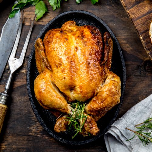 A flat lay pic of a roasted chicken with rosemary served on black plate with sauces on wooden table