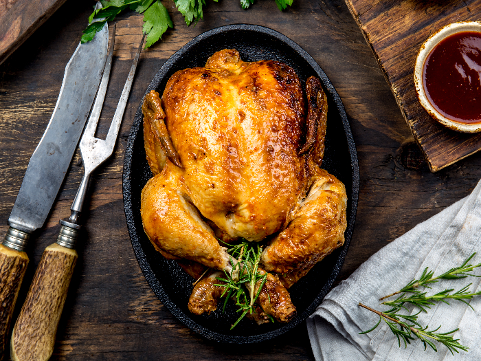 A flat lay pic of a roasted chicken with rosemary served on black plate with sauces on wooden table