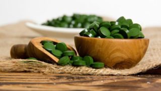 A wooden bowl filled with green pills next to a spoon full of the same atop a jute mat