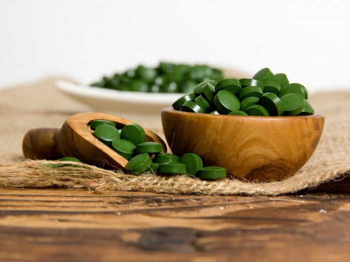 A wooden bowl filled with green pills next to a spoon full of the same atop a jute mat