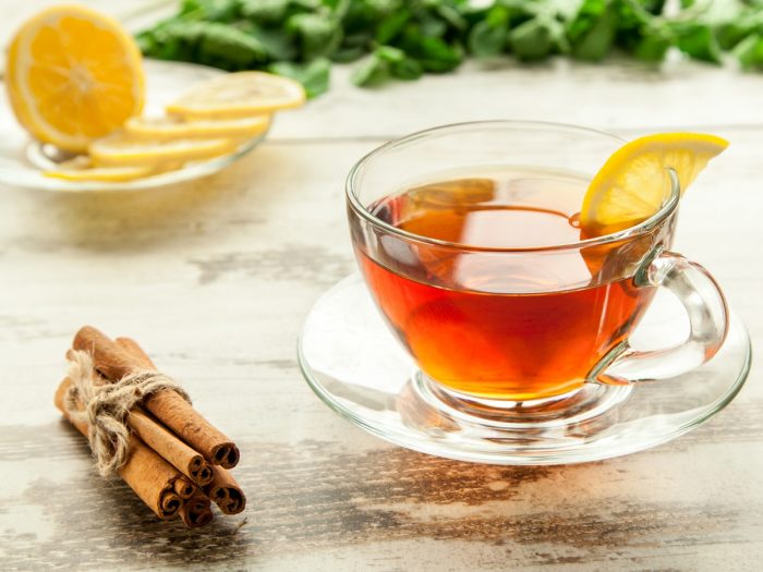 A cup of fresh cinnamon tea kept atop a table, next to a bunch of cinnamon sticks and a plate of lemon slices