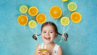A smiling girl holding a glass of juice with flying citrus halves tied to her pigtails, standing against a blue background.