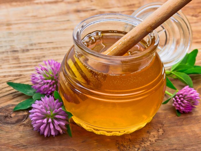 A jar of clover honey, a wooden dipper, and red clovers on a wooden table