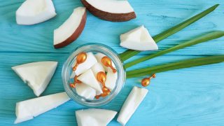 A bowl of sliced coconuts and coconut capsules surrounded by sliced coconuts and leaves on a blue wooden background