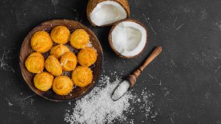 Coconut flour biscuits (cookies) on a wooden plate, kept beside ingredients like fresh chopped coconut and coconut chips, atop a dark black concrete table background.