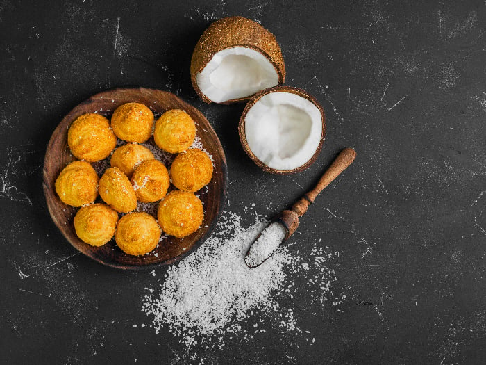 Coconut flour biscuits (cookies) on a wooden plate, kept beside ingredients like fresh chopped coconut and coconut chips, atop a dark black concrete table background.