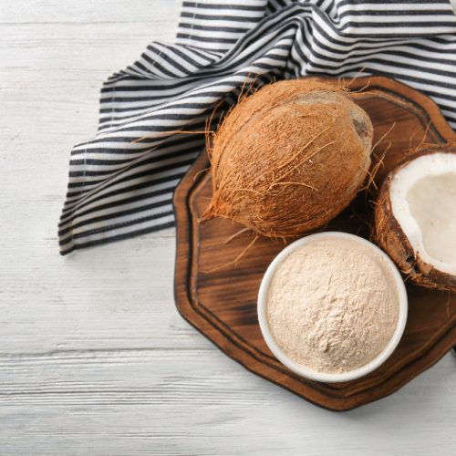 Coconuts and a bowl of coconut flour on a wooden board with a black & white striped napkin.