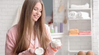 A woman with long hair in the bathroom holding a coconut and a hair mask bottle