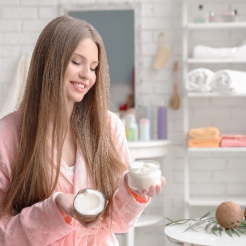 A woman with long hair in the bathroom holding a coconut and a hair mask bottle