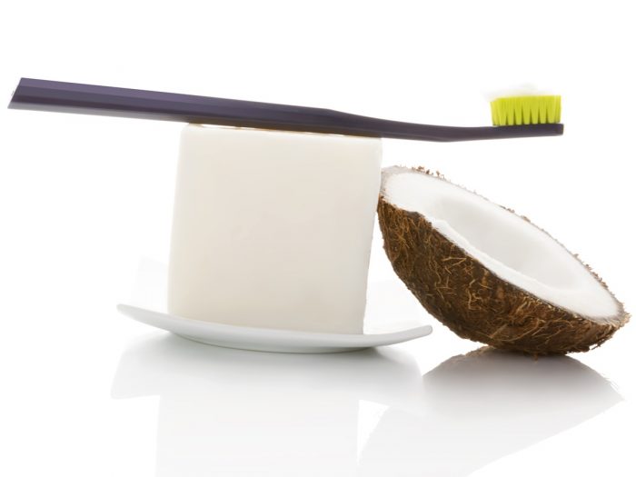 A coconut shell placed alongside a white bar with a toothbrush placed on top of a white dish with a white background.