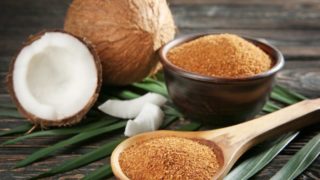 A wooden spoon and a bowl of coconut sugar with a whole and halved coconuts on a wooden table