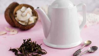 A picture of a white kettle containing coconut tea, placed in front of dried coconut pieces against a pink background