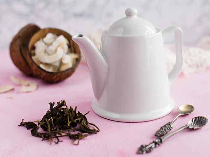 A picture of a white kettle containing coconut tea, placed in front of dried coconut pieces against a pink background