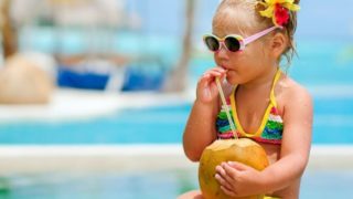 A small girl wearing a colorful swimsuit drinking coconut water near a swimming pool