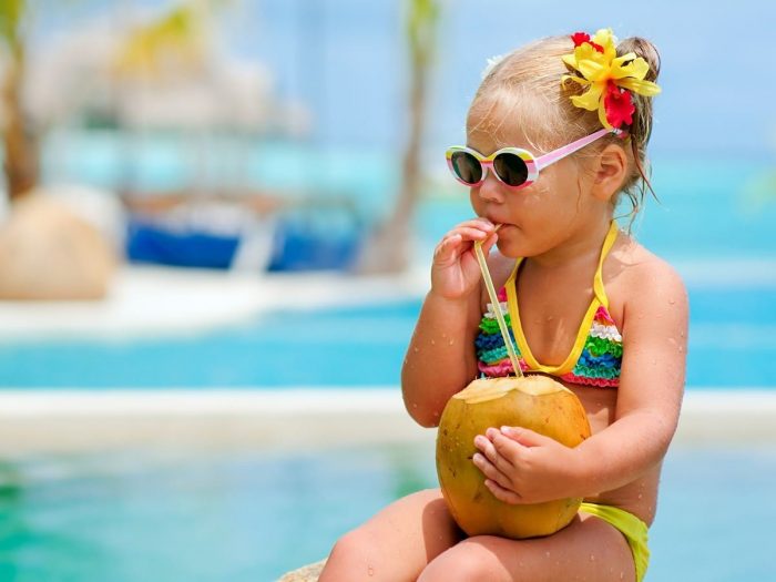 A small girl wearing a colorful swimsuit drinking coconut water near a swimming pool