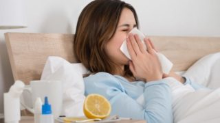 A woman sneezing into a tissue paper with medicines and lemon on the side table