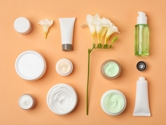 Bottles, jars, and tubs containing skincare formulations along with flowers on a peach background.