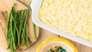 Close up shot of casserole dish with of unbaked creamy cheese corn casserole kept on a wooden table, next to bowls of cheese and asparagus