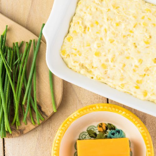 Close up shot of casserole dish with of unbaked creamy cheese corn casserole kept on a wooden table, next to bowls of cheese and asparagus