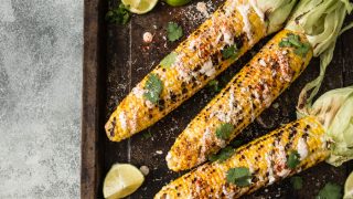 A flat lay picture of grilled corn cobs with sauce, coriander, lime, paprika and cheese.