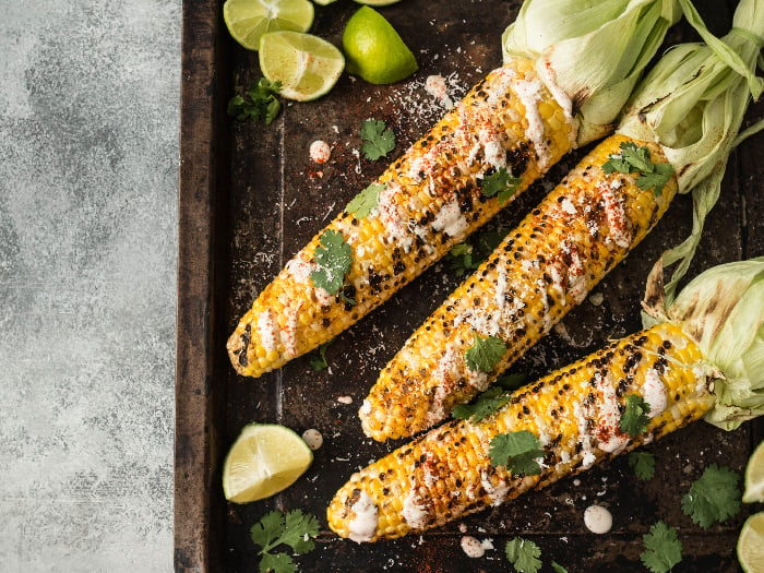 A flat lay picture of grilled corn cobs with sauce, coriander, lime, paprika and cheese.