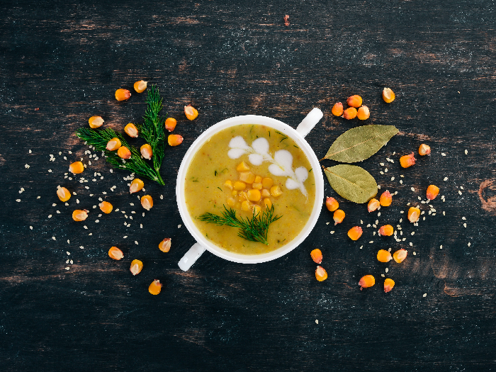 A flat lay picture of healthy corn soup with fresh vegetables in a bow kept atop a black wooden background.