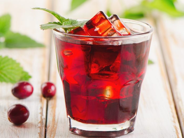 A glass of red-colored liquid in a glass-topped with ice and a sprig of mint on a wooden surface