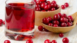 A wooden bowl and a spoon full of cranberries with a glass of cranberry juice on a wooden surface.