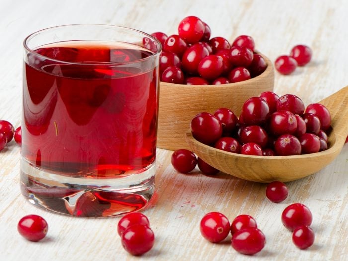 A wooden bowl and a spoon full of cranberries with a glass of cranberry juice on a wooden surface.