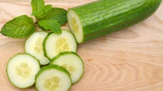 Freshly sliced cucumber with the whole vegetable and a mint leaf on a wooden cutting board
