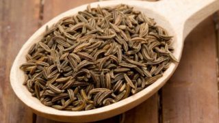 A  wooden spoon full of cumin placed on a wooden surface.