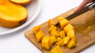 A plate of sliced mango next to a wooden chopping board with small pieces of mango being cut with a knife